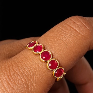 Op de afbeelding: Een gouden ring met vijf ovaal geslepen, helderrode edelstenen. De edelstenen zijn gezet in afzonderlijke gouden bezels, waardoor een doorlopende lijn ontstaat. De ring wordt getoond tegen een donkere achtergrond.