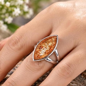 Puede incluir: Un anillo de tono plateado con un engaste en forma de marquesa y una incrustación de piedra texturizada de color ámbar. La piedra muestra un aspecto brillante y reluciente en tonos naranja y dorado, sostenida por una banda de metal pulido dividida.