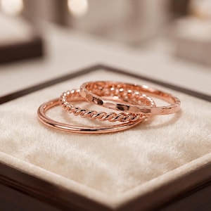 May include: A set of three rose gold rings featuring distinct textures. One ring has a smooth, polished surface, the second displays a twisted rope design, and the third ring has a hammered, faceted finish. The rings are stacked together.