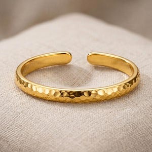 Könnte beinhalten: Ein goldfarbenes offenes Armband mit gehämmertem Muster. Das Armband besteht aus einem glänzenden, metallischen Material und hat ein schlichtes, elegantes Design. Das Armband wird auf einem neutralfarbenen Stoff gezeigt.