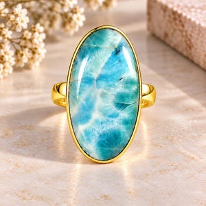 May include: A gold ring featuring a large, oval-shaped, polished larimar gemstone. The stone displays a marbled pattern of light blue, turquoise, and white hues with subtle dark inclusions, securely set within a smooth, polished gold-tone metal bezel.