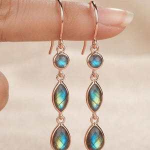 Puede incluir: Un par de pendientes colgantes de oro rosa con tres piedras de labradorita alineadas verticalmente. Cada pendiente consta de una pequeña piedra redonda en la parte superior, una piedra en forma de marquesa en el centro y una gota en la base.