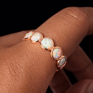May include: A rose gold ring with five oval opals. The opals show a play of colour, with flashes of iridescent hues. The ring is set against a dark background, highlighting the jewellery's design.
