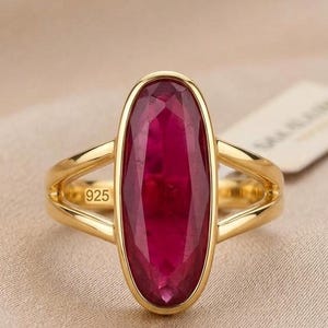 Può includere: Anello in oro con una grande gemma di rubino ovale. Il rubino è di un rosso intenso ed è incastonato in una lunetta d'oro. L'anello è timbrato con il numero "925". Presentato su sfondo neutro.