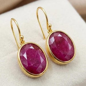 May include: A pair of gold earrings featuring oval, faceted, deep red gemstones. The gemstones are set in a gold bezel and suspended from gold ear wires. The earrings are presented on a white surface.