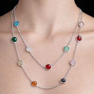 Può includere: Una collana d'argento con due fili, ciascuno ornato da pietre preziose rotonde e colorate. Le pietre includono blu, verde, bianco, giallo, rosso, rosa, arancione e blu scuro. La collana è presentata su sfondo nero.