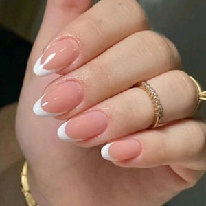 Pode incluir: Close-up de uma mão com unhas ovais pintadas com um esmalte nude e uma ponta branca francesa.