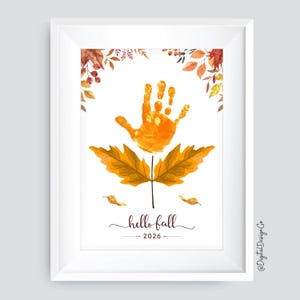 Könnte beinhalten: Ein gerahmter Druck mit einem zentralen orangefarbenen Handabdruck über zwei großen braunen Herbstblättern. Das Design enthält aquarellartige Blätter in den oberen Ecken und den Text "hello fall 2026" in einer dunkelbraunen Schreibschrift.