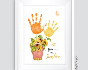 May include: A white framed print with two orange and yellow handprints in the shape of sunflowers. The handprints are in a pink pot with a bouquet of sunflowers. The text "You are our Sunshine" is below the pot.