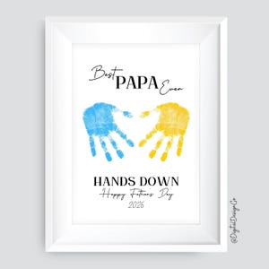 以下が含まれることがあります： 「Best PAPA Ever」の文字と青と黄色の手形が描かれた白いフレームのプリント。手形の下には「HANDS DOWN Happy Father's Day 2026」と書かれています。父の日のギフトに最適です。