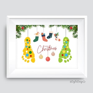 May include: Framed Christmas art print featuring two footprint Christmas trees, colorful ornaments, and hanging stockings. The words "Merry Christmas" are written in a festive font. The print is decorated with evergreen branches.