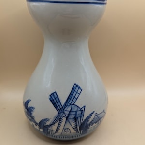 Puede incluir: Un jarrón de cerámica blanca con un diseño de molino de viento azul y blanco. El jarrón tiene una boca ancha y una base estrecha.