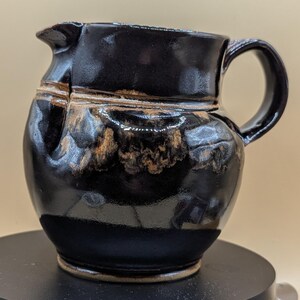 May include: A handmade black ceramic jug with a handle and spout. The jug has a glossy finish with brown and tan accents. The ceramic jug is a unique piece of pottery.