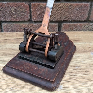 Op de afbeelding: Een antieke perforator met een houten basis en koperen en stalen componenten. De handgreep is zilverkleurig en het ponsmechanisme is een mix van koper en zwart metaal. De houten basis is donkerbruin.