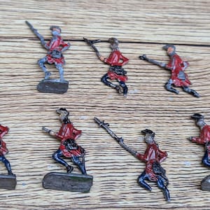 Puede incluir: Colección de siete soldaditos de juguete en miniatura. Cada figura lleva una casaca roja, pantalones negros y una base gris. Algunos soldados sostienen armas. Las figuras están dispuestas sobre una superficie de madera.