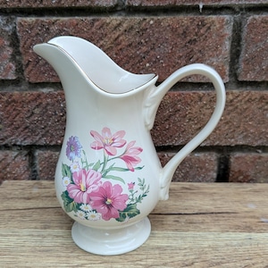 Puede incluir: Jarra de cerámica color crema con un diseño floral de flores rosas, moradas y blancas. La jarra tiene un borde dorado, un pico curvo y un asa. Se asienta sobre una base de pedestal.
