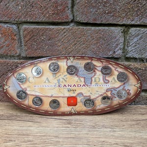 Puede incluir: Un expositor ovalado de madera con monedas canadienses del Milenio. El expositor tiene un diseño de mapa con las palabras "CANADA MILLENAIRE 1999" y un cuadrado rojo con una hoja de arce. Las monedas están dispuestas alrededor del mapa.
