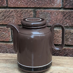 Könnte beinhalten: Eine braune Keramik-Teekanne mit Deckel und Henkel. Die Teekanne hat ein weißes und schwarzes Band um den Boden. Der Hintergrund ist eine Ziegelmauer und eine Holzoberfläche.