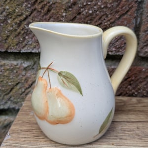 Può includere: Una brocca in ceramica color crema con finitura maculata, caratterizzata da un disegno dipinto di due pere arancione chiaro con foglie verdi sul davanti. La brocca presenta un manico curvo e un ampio beccuccio con una superficie smaltata.