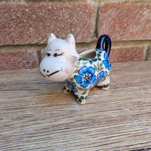 Op de afbeelding: Een keramieken melkkannetje in de vorm van een koe, met een witte kop voorzien van geschilderde zwarte ogen, wimpers en een glimlach. Het lichaam is versierd met een blauw bloemmotief op een crèmekleurige achtergrond en een donkerblauw handvat.