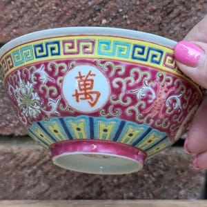 May include: A small, decorative porcelain bowl with a vibrant design. The bowl features a red background with white floral patterns and a central circular emblem with a Chinese character. The rim has a yellow and blue geometric border.