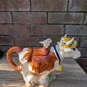 May include: A ceramic teapot shaped like a brown and white cow with a small bird perched on its back. The cow has a yellow and blue hat and a bell around its neck.
