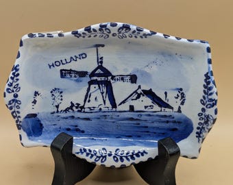 May include: A small, white ceramic dish with a blue and white windmill scene. The dish has a scalloped edge and is decorated with a blue and white floral pattern. The dish says "HOLLAND" in blue lettering. The dish is on a black stand.