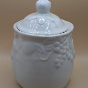 Op de afbeelding: Witte suikerpot van keramiek met een deksel. De pot is versierd met een druivenranken-ontwerp.