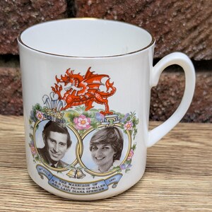 May include: White ceramic mug with a gold rim, featuring portraits of Prince Charles and Lady Diana Spencer, with a red dragon emblem. The mug commemorates their marriage in July 1981.