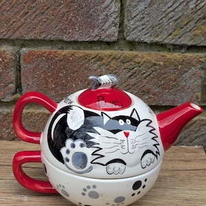 May include: A novelty white teapot with a red handle and spout, decorated with a black and white cat motif. The lid is red and has a fish-shaped handle. The teapot sits on a matching cup, decorated with paw prints. The word "Paws" is written on the lid.