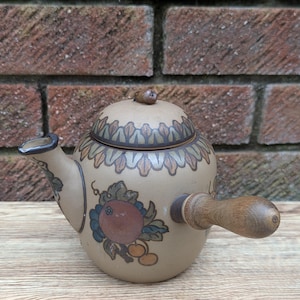 Pode incluir: Uma chaleira de cerâmica marrom com uma alça de madeira e um design floral. A chaleira tem uma tampa com uma pequena maçaneta no topo. A chaleira está sentada em uma superfície de madeira.