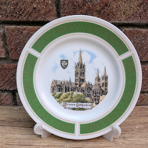 May include: A white ceramic decorative plate featuring a central illustration of Truro Cathedral with a small crest above it. The plate has a wide green border with a repeating scroll pattern and gold trim accents along the inner and outer edges.