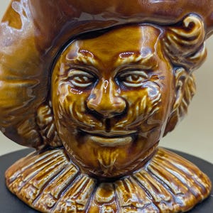 May include: A brown ceramic figurine of a smiling man wearing a hat and a ruffled collar. The figurine has a glossy finish and is set against a dark background. The face has detailed features.