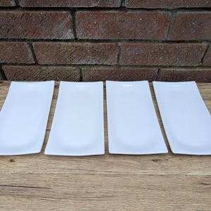 May include: Four white rectangular ceramic serving dishes, arranged in a row on a wooden surface.