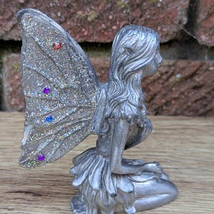 Op de afbeelding: Zilverkleurig feeënbeeldje met glittervleugels. De fee knielt, met gevouwen handen. De vleugels zijn versierd met gekleurde edelstenen. Het beeldje staat op een houten oppervlak.