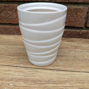 May include: A white ceramic plant pot with a textured, wave-like pattern. The small, cylindrical pot is on a wooden surface, with a brick wall behind it. The pot is suitable for small plants and succulents.