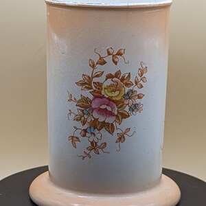 May include: A cylindrical ceramic vase with a floral design. The vase has a light peach base and rim, with a white body featuring a bouquet of pink, yellow, and blue flowers. The vase is approximately 13 cm tall.