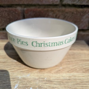 May include: A cream-coloured ceramic mixing bowl with a tapered shape. The exterior rim features green text that reads Mince Pies Christmas Cakes. The bowl has a smooth, glossy finish and a wide opening, designed for kitchen use and food preparation.