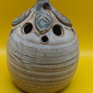 Könnte beinhalten: Ein weißer Keramiktopf mit brauner und grüner Glasur. Der Topf hat eine abgerundete Form und mehrere Löcher oben. Es ist ein dekorativer Topf für Pflanzen oder Blumen.