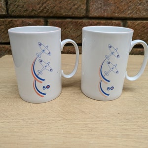 May include: A pair of white ceramic mugs with handles. Each mug has a design of two aeroplanes with red, white, and blue curved lines, and the number "50". The mugs are on a light-coloured wooden surface.
