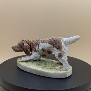 May include: A ceramic dog figurine, mainly white with brown patches and a brown head, wearing an orange collar. The dog is posed on a small, oval base with a grassy texture. The figurine is on a black rotating platform.