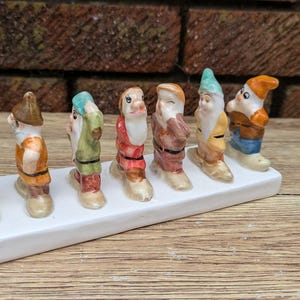 Peut inclure: Ensemble de sept figurines de nains en céramique, chacune avec une tenue et un chapeau uniques, disposées sur une base rectangulaire blanche. Les nains sont peints dans diverses couleurs, dont le jaune, l'orange, le vert et le marron.