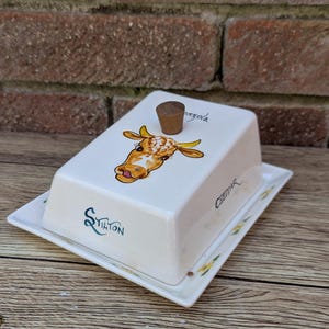 Könnte beinhalten: Eine weiße Keramik-Butterdose mit einem Kuh-Design und einem Holzgriff auf dem Deckel. Die Dose hat einen kleinen Teller und die Worte "Stilton" und "Gorgonzola" sind auf der Seite aufgedruckt.