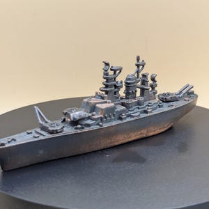 Puede incluir: Maqueta detallada de un acorazado en miniatura en tonos grises y marrones. El modelo presenta intrincados detalles de la estructura del barco, incluyendo torretas, mástiles y otros componentes. El barco se exhibe sobre una superficie oscura.