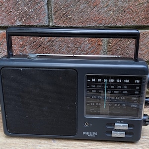 May include: A dark grey Philips portable radio featuring a black speaker grille, a top carrying handle, and a rectangular tuning dial with frequency scales for FM, LW, MW, SW1, and SW2. Two silver sliding switches are located below the dial.