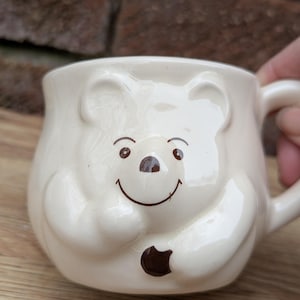 Może przedstawiać: Biały kubek ceramiczny z brązową twarzą niedźwiedzia i brązowym śladem łapy po bokach. Kubek ma zaokrąglony kształt i uchwyt.