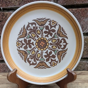 May include: A white ceramic plate with a brown and gold floral design. The plate has a brown and gold border and a central pattern of brown leaves and flowers with accents of orange and teal. The plate is displayed on a wooden stand.