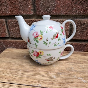 May include: A white ceramic tea-for-one set consisting of a teapot stacked on top of a matching teacup. The set features a repeating pattern of small red-breasted birds, pink and yellow roses, green leaves, and small blue flowers.