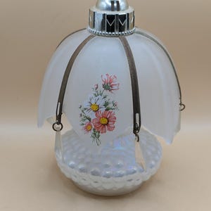 Può includere: Una lampada in vetro bianco con un motivo floreale e una gabbia di metallo. La lampada ha un paralume in vetro bianco smerigliato e una base bianca iridescente.