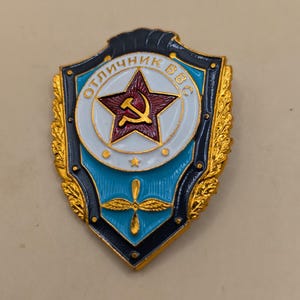 May include: A vintage military badge in a shield shape. The badge features a red star with a hammer and sickle emblem at the centre, surrounded by text in a circular design. The badge has gold, blue, and white accents.